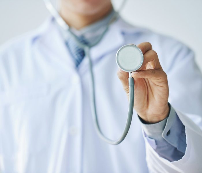 Doctor with stethoscope Obraz przedstawia osobę ubraną w biały fartuch laboratoryjny, trzymającą stetoskop uniesiony w kierunku obiektywu. Ujęcie skupia się na stetoskopie, co podkreśla jego znaczenie jako symbolu zawodu medycznego. Postać prawdopodobnie reprezentuje lekarza lub pielęgniarkę. Tło jest neutralne, co pozwala skupić uwagę na głównym elemencie – narzędziu diagnostycznym.