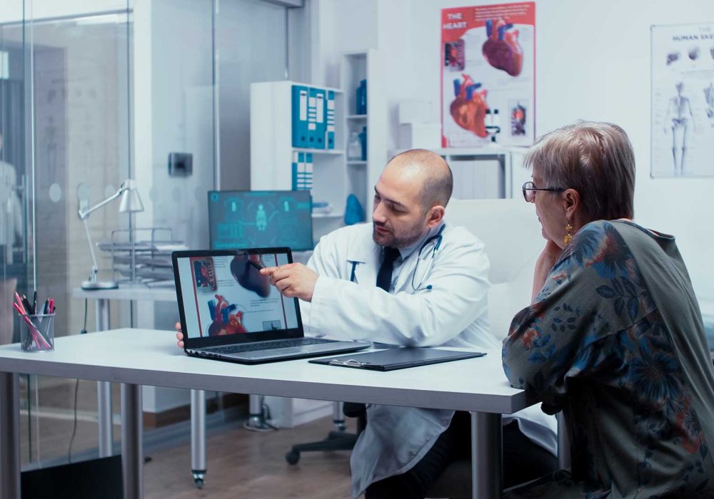 explaining-possible-heart-disease-XG7ETS8.jpg Obraz przedstawia scenę w gabinecie lekarskim, gdzie lekarz omawia z pacjentem obraz serca wyświetlony na ekranie laptopa. Lekarz wskazuje na grafikę, co sugeruje wyjaśnianie diagnozy lub planu leczenia. W tle widoczne są plakaty medyczne – jeden przedstawia anatomię serca, drugi szkielety ludzkie. W pomieszczeniu znajdują się również elementy wyposażenia medycznego, takie jak stół do badań i różne narzędzia diagnostyczne. Całość tworzy profesjonalne i edukacyjne środowisko opieki zdrowotnej.