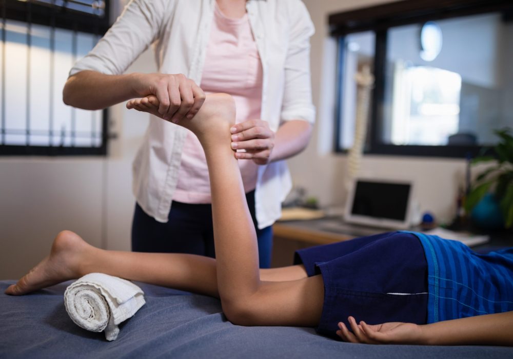 Low section of boy receiving foot massage from young female therapist W pomieszczeniu przypominającym gabinet fizjoterapeutyczny lub salę rehabilitacyjną widoczna jest scena, w której terapeuta wykonuje rozciąganie kończyny dolnej pacjenta leżącego na stole zabiegowym. Pacjent ułożony jest na plecach, z jedną nogą zgiętą w kolanie i podpartą pod kostką zwiniętym ręcznikiem, co zapewnia stabilizację i komfort podczas zabiegu. Terapeuta, stojąc z boku, delikatnie trzyma stopę i podudzie pacjenta, wykonując kontrolowany ruch rozciągający. W tle widoczne są elementy wyposażenia medycznego, takie jak szafki, sprzęt terapeutyczny oraz neutralne ściany, co podkreśla profesjonalny charakter miejsca. Cała scena ukazuje precyzyjne i uważne podejście do pracy z ciałem pacjenta w kontekście rehabilitacji lub terapii manualnej.