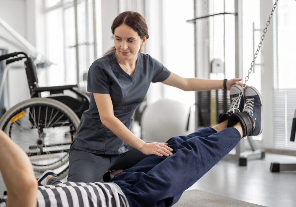 W przestrzeni terapeutycznej widoczna jest scena, w której fizjoterapeuta wspiera pacjenta podczas ćwiczeń kończyn dolnych. Pacjent leży na macie na podłodze, unosząc obie nogi w powietrzu, podczas gdy terapeuta, klęcząc obok, stabilizuje i kontroluje ruchy nóg, zapewniając prawidłowe wykonanie ćwiczenia. W tle znajdują się różnorodne elementy wyposażenia rehabilitacyjnego, w tym wózek inwalidzki oraz sprzęt do ćwiczeń, co wskazuje na profesjonalne środowisko wspierające proces powrotu do sprawności. Całość kompozycji podkreśla zaangażowanie terapeuty i aktywny udział pacjenta w terapii ruchowej.